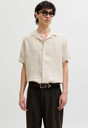 Młody mężczyzna w czarnych okularach przeciwsłonecznych, beżowej koszuli z krótkim rękawem zapinanej na guziki, czarnych spodniach i czarnym pasku ze srebrną klamrą.