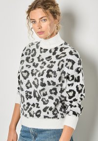 Biała sweter z czarnym wzorem w panterę, ozdobiony cekinami i z wysokim kołnierzem. Żebrowane mankiety i dół. Noszony na niebieskich dżinsach.