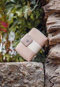 Růžová a krémová kožená peněženka s uzávěrem na knoflík a embosovaným logem "VUCH", ležící na kamenném povrchu s zelení v pozadí.