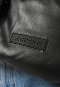 Bolso de cuero sintético negro con un acabado texturizado que presenta una etiqueta rectangular negra grabada con "SIXTHJUNE." Diseño sencillo sin patrones.