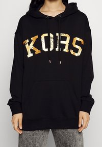 Felpa nera con cappuccio e tasca frontale, dotata di un logo "KORS" in oro rovinato. Realizzata in tessuto morbido, con coulisse al cappuccio e polsini a costine.