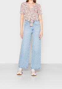 Donna che indossa una blusa legata davanti con motivo paisley rosa, jeans a vita alta e a gamba larga di colore blu chiaro e sneakers bianche con suole arancioni.