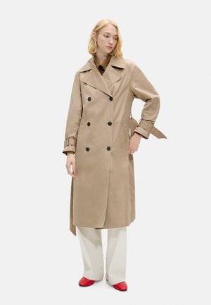Blondynka w beżowym płaszczu dwurzędowym, białe spodnie o szerokich nogawkach i czerwone buty, stojąca z jedną ręką w kieszeni.