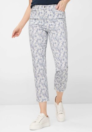 Person iført cropped bukser med blå og gråt diamantmønster og hvide sneakers, stående mod en ensfarvet baggrund.