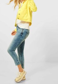 Gelbe Jeansjacke, weißes Hemd, hellblaue enge Jeans und beigefarbene Sneakers mit cremefarbenen Akzenten, mit einem leichten Knöchelschnitt und seitlichem Reißverschluss.