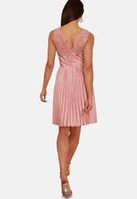 Ljusrosa klänning med plisserad kjol, blommig spetsdetaljer på axelbanden och en V-formad ryggdesign. Texturerat, lättviktsmaterial.