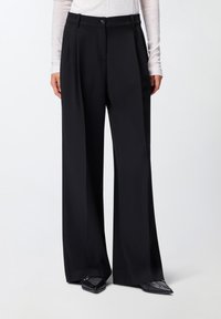 Zwarte wijde broek van soepel materiaal met een hoge taille, een knoopsluiting en voorplooien. Gemaakt in combinatie met puntige zwarte schoenen.