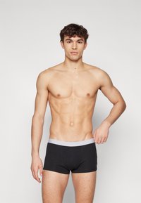 Svarte boksershorts med en grå elastisk midje, laget av et glatt, stretchy stoff. Modellen står med et nøytralt uttrykk.