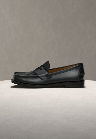 Juodas odinis batelis („loafer“) su austiniu paviršiumi viršuje, plokščia padu ir žemu kulnu, eksponuojamas ant šviesaus paviršiaus neutraliame fone.