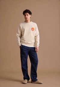 Sweat-shirt crème avec logo orange sur la poitrine, associé à un pantalon en denim bleu foncé. Le mannequin porte des chaussures beige à enfiler, debout devant un fond neutre.