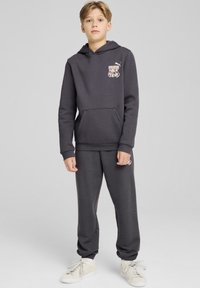 Sudadera gris con capucha y bolsillo frontal, con un logo. Combinada con pantalones de chándal grises a juego. Zapatillas blancas completan el look.