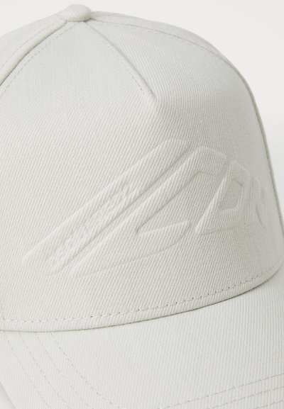 Lys beige kasket med et struktureret stof, der har et hævet logodesign på forsiden og en buet skygge. Syning detaljeret på kanterne.