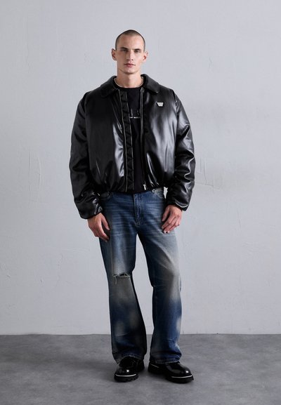 Homme portant une veste en cuir noir, une chemise noire, un jean bleu déchiré et des chaussures noires, se tenant contre un mur gris uni.