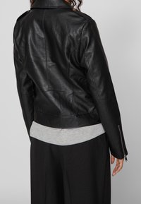 Svart läderjacka med en kortad design, synliga sömdetaljer och dragkedja vid ärmarna. Bärs över en grå sweatshirts och svarta byxor.