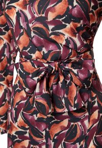 Gros plan sur une ceinture en tissu nouée avec un imprimé floral abstrait dans des tons rouille, violet, noir et beige sur un vêtement à manches longues.