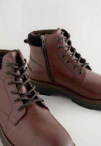 Braune Leder-Ankelstiefel mit strukturiertem Finish, ausgestattet mit einer Schnürung vorne und einem seitlichen Reißverschluss. Gummisohle für guten Halt.
