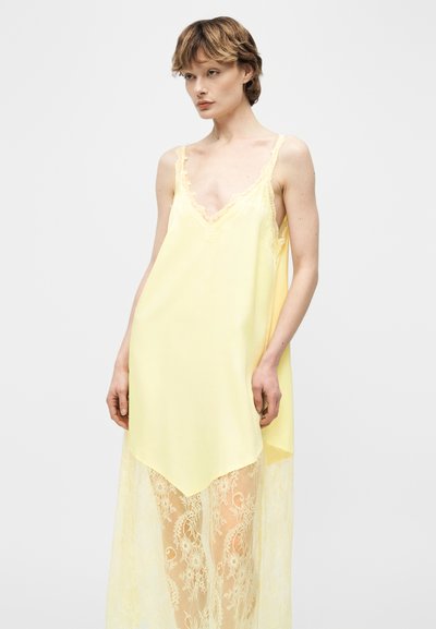 Γυναίκα που φοράει κίτρινο φόρεμα χωρίς μανίκια με δαντελένιες λεπτομέρειες, όρθια μπροστά σε ένα απλό ανοιχτό γκρι φόντο.