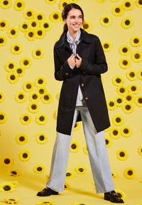 Schwarzer Trenchcoat mit großen Knöpfen, hellblaue weitgeschnittene Jeans und schwarze Slipper, vor einem gelben Hintergrund mit Sonnenblumen.