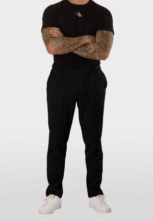 Hombre de pie con los brazos cruzados, llevando mangas negras tatuadas, camiseta negra, pantalones deportivos negros y zapatillas blancas sobre fondo blanco.