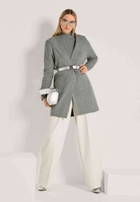 Vrouw in een oversized grijze gebreide cardigan met riem, witte wijdlopende broek, zilveren tas en ronde zonnebril, staande tegen een lichte achtergrond.