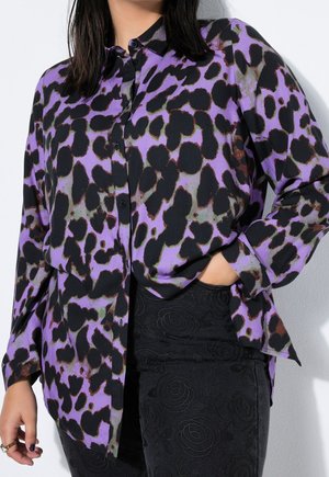 Femme portant une chemise boutonnée à motif abstrait violet et noir avec la main dans la poche d’un pantalon noir à texture florale.