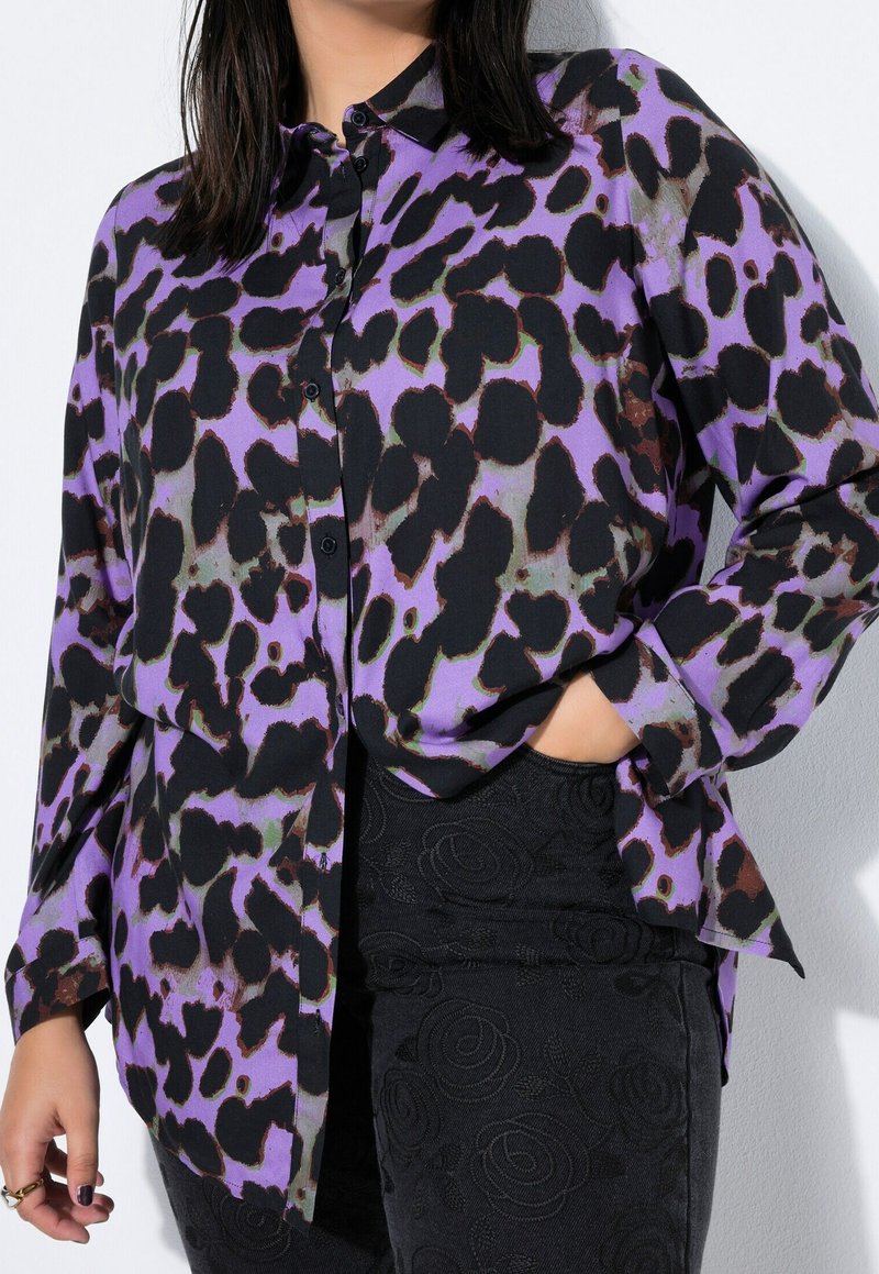 Femme portant une chemise boutonnée à motif abstrait violet et noir avec la main dans la poche d’un pantalon noir à texture florale.
