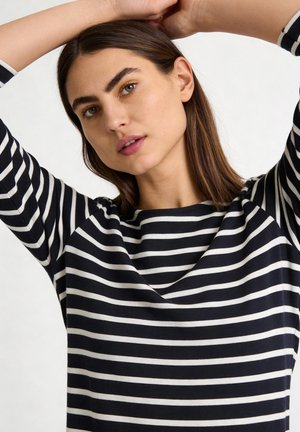 Young woman with brown hair wearing a black and white striped long-sleeve shirt, arms raised, looking slightly to the side.