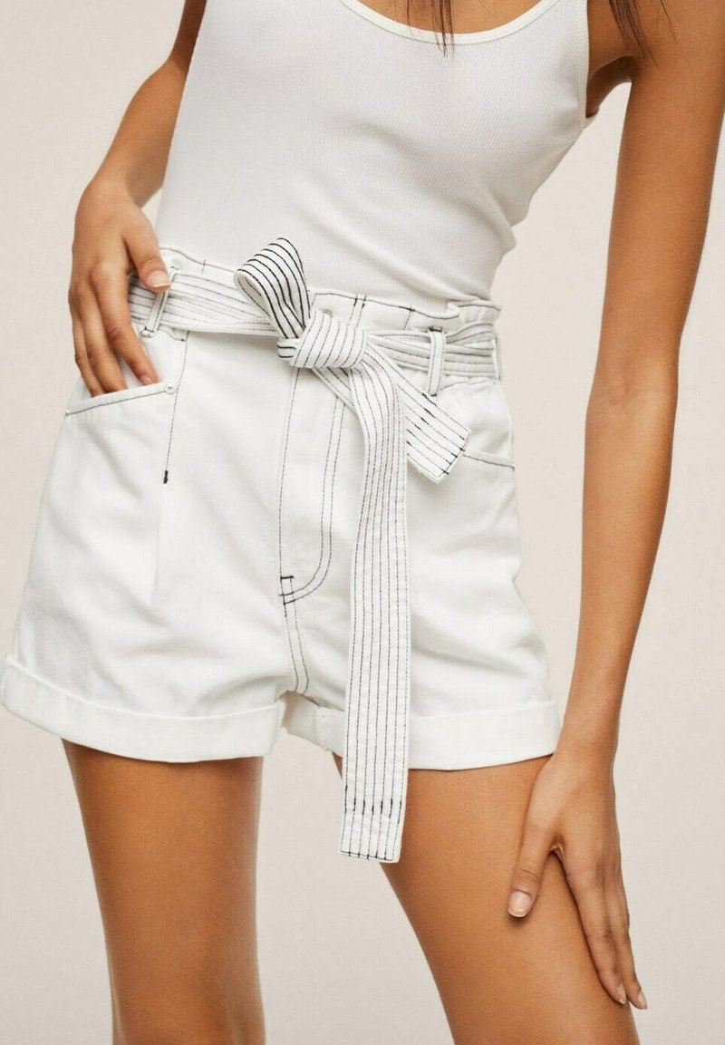 Femme portant un short blanc taille haute avec des revers roulés et une ceinture large en tissu rayé nouée en nœud, associée à un haut blanc sans manches.