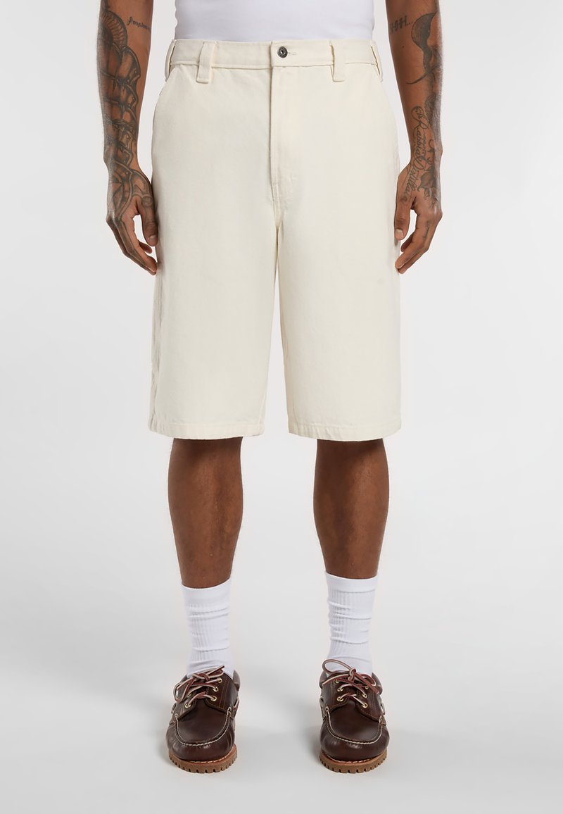 Pantaloni corti in cotone beige con vestibilità dritta, lunghezza al ginocchio e chiusura anteriore con bottone. Indossati con calzini bianchi e scarpe marroni.