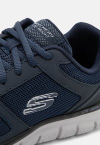Marineblaues Netz-Sport-Schuhwerk mit strukturierten synthetischen Akzenten, ausgestattet mit einem silbernen Logo, schwarzer Sohle und verstärkten Ösen für die Schnürsenkel.