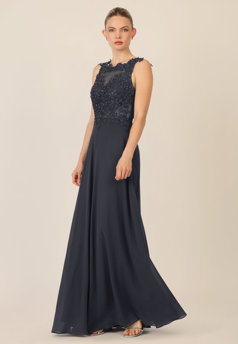 Marineblaue bodenlange Kleid mit einem transparenten, bestickten Oberteil und kurzen Ärmeln, glattem, fließendem Rock und schmalen Trägern. Sichtbare silberne Schuhe.