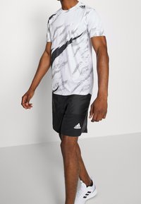 Camiseta deportiva blanca para hombre con un patrón abstracto negro, combinada con pantalones cortos negros. Incluye el logo de Adidas en blanco en los pantalones cortos y zapatos negros.