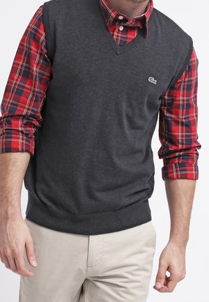 Mann trägt rotes kariertes Hemd mit hochgekrempelten Ärmeln unter dunkelgrauem ärmellosem Pullover und beigen Hosen.