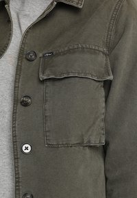 Gros plan sur une chemise boutonnée vert olive avec une poche à rabat et des boutons en métal portée sur un t-shirt gris clair.