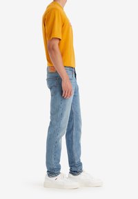 Gul bomullströja med korta ärmar, blå denimjeans och vita sneakers. T-shirten har en logotryck. Jeansen har rak passform.