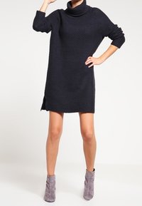 Robe pull en tricot bleu marine avec un col cheminée, des manches longues et des fentes sur les côtés, assortie à des bottines grises à la cheville. Tissu texturé avec une coupe ample.