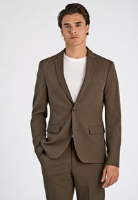 Mann in einem olivgrünen Anzug mit schwarzen Knöpfen über einem weißen T-Shirt, der mit einer Hand in der Tasche vor einem schlichten weißen Hintergrund steht.