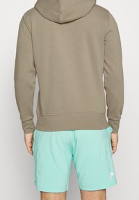 Muž v taupe mikině a světlých tyrkysových sportovních šortkách, zobrazený zezadu na jednobarevném bílém pozadí.