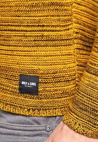 Žlutý pletený svetr s horizontálními pruhy, s žebrovaným lemem a manžetami. Na spodní straně přišitá etiketa. Světle šedé pozadí.