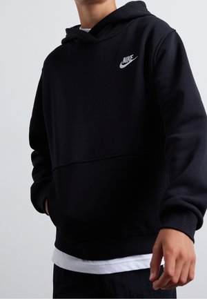 Osoba ubrana w czarną bluzę z kapturem Nike z rękami w kieszeniach, na której widać wystający biały brzeg koszuli, na jasnym tle.