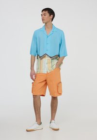 Camicetta blu a maniche corte con bottoni e orlo a fantasia colorata, abbinata a pantaloni cargo arancioni con tasche laterali. Sneakers bianche.