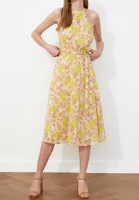 Robe midi florale en pêche avec des fleurs jaunes et orange, design à encolure halter, taille cintrée avec une ceinture à nouer, jupe fluide, tissu texturé.