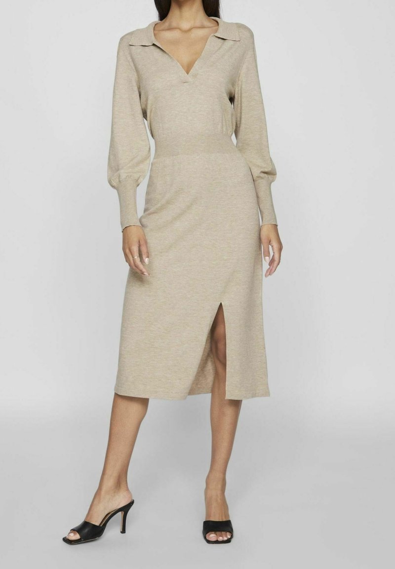 Femme portant une robe midi beige à manches longues avec col et fente frontale, associée à des mules noires à talons ouvertes à l'avant.