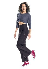 Grauer, verkürzter Oberteil mit Knoten-Detail, hochgeschnittene schwarze Hose und kastanienbraune Sneakers. Glattes Material, lockere Passform und kontrastierende Farben.