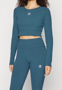 Žena v odpovídajícím námořnickém modrém vroubkovaném dlouhém rukávu od Adidas a legínách s vysokým pasem, na hrudi a stehně s bílými logy Adidas.