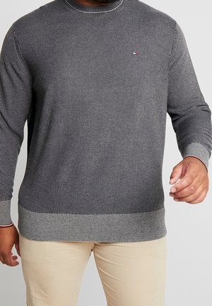 Man wearing grey textured sweater with ribbed cuffs and hem, paired with beige pants, against plain light background.