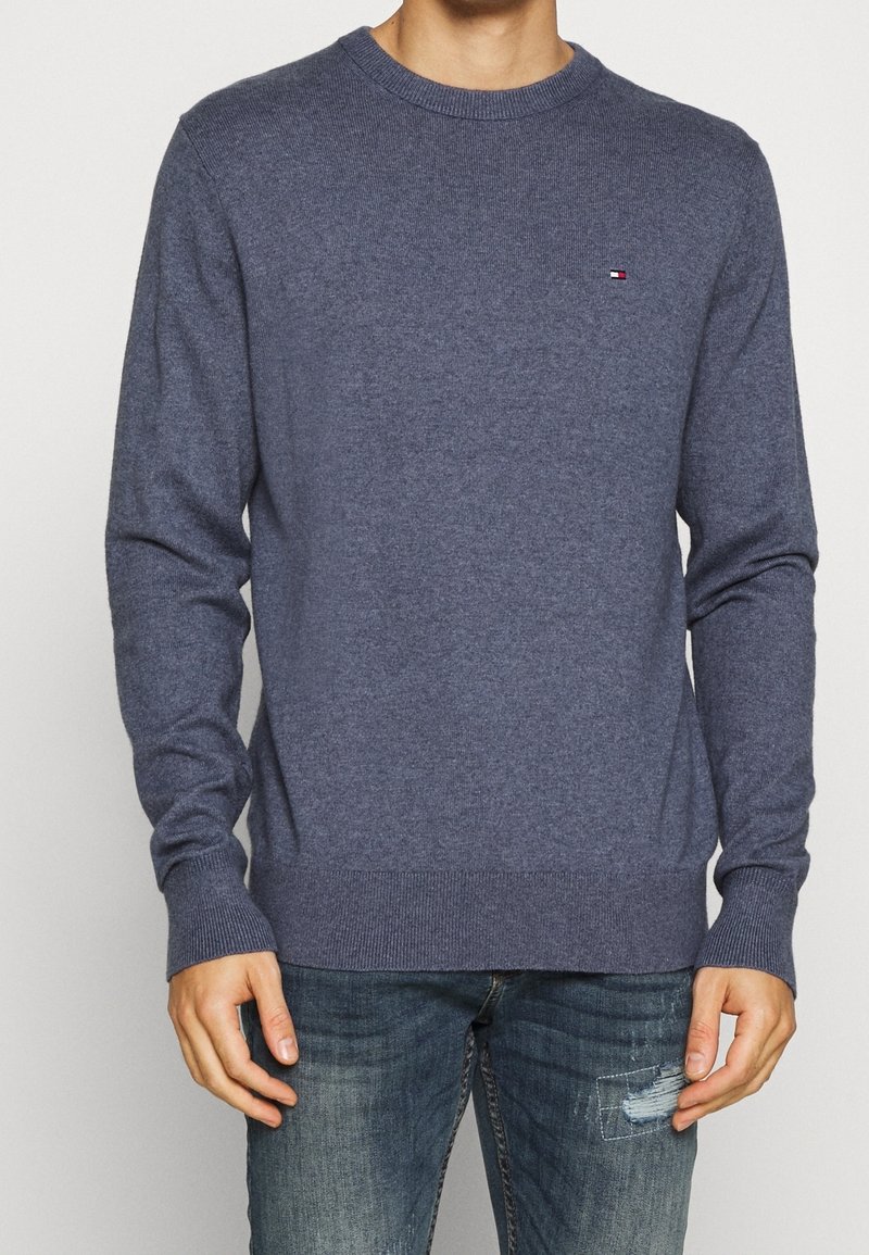 Homme portant un pull ras-du-cou bleu uni avec des poignets côtelés et un petit logo sur la poitrine, assorti à un jean bleu déchiré.