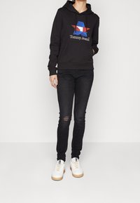 Černá mikina s kapucí a červeným a modrým grafickým designem hvězdy a logem "Tommy Jeans", spárovaná s černými skinny džínami a bílými teniskami.