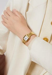 Montre-bracelet dorée avec un cadran blanc carré, des index horaires bleus et un bracelet en métal doré texturé, portée sur une manche de couleur claire.
