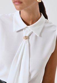 Vrouw in een mouwloze witte blouse met een kraag en een geplooide voorkant die wordt vastgehouden door een gouden ring sluiting.